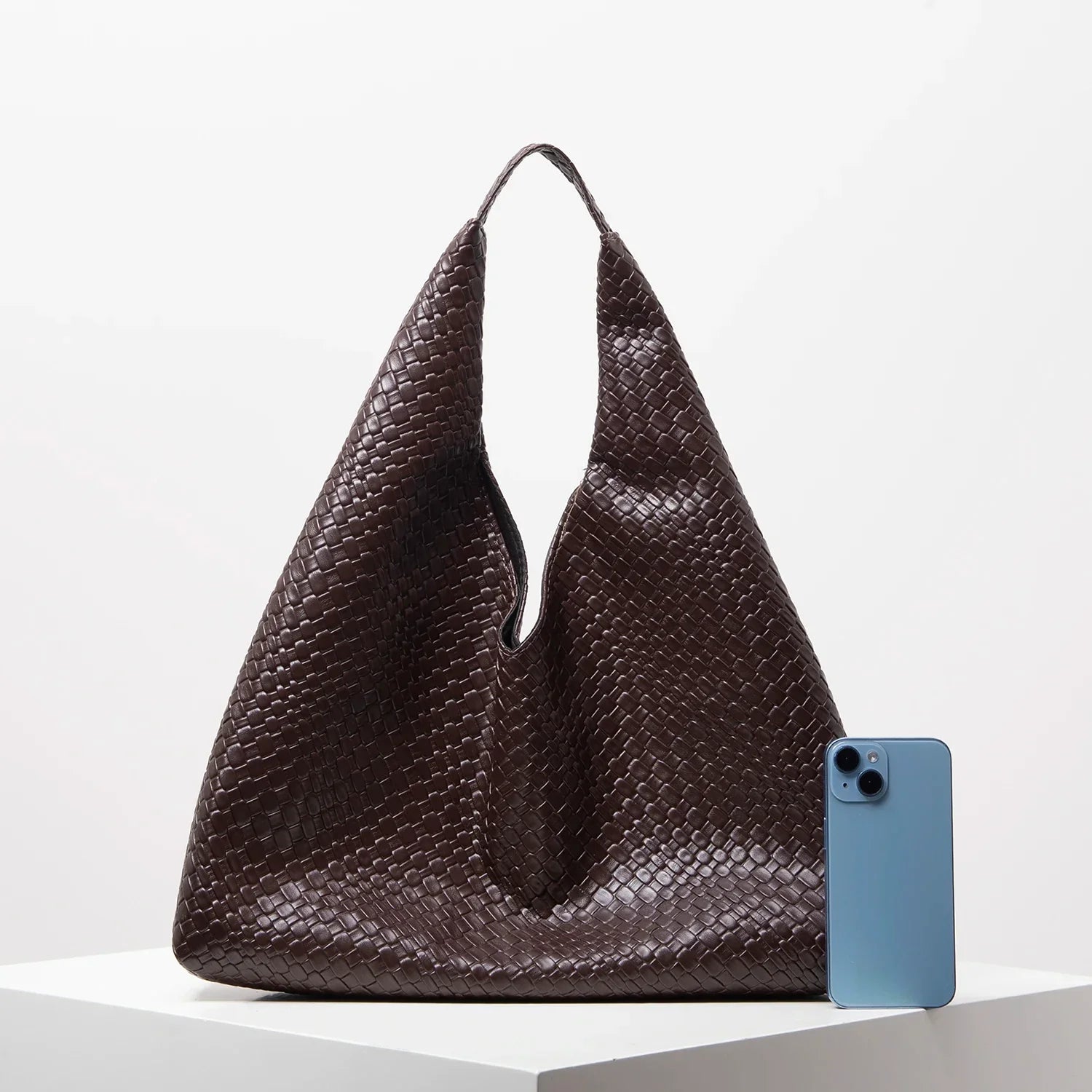 Bolsa grande marrom de couro trançado e celular azul sobre superfície branca